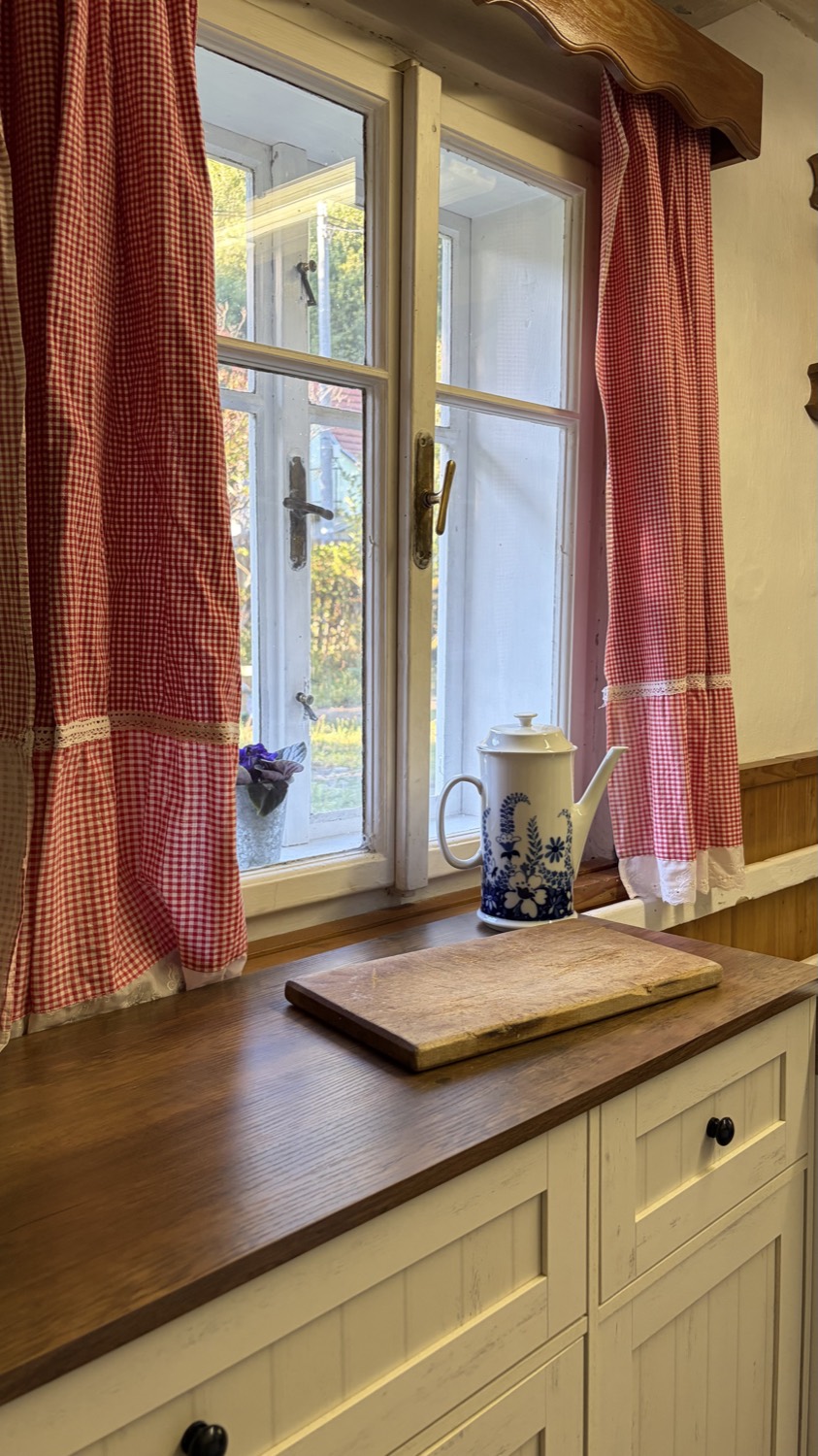 Kuchyňské okno s kytkovanými záclonami, modrobílá porcelánová konvice na parapetu a dřevěné prkénko